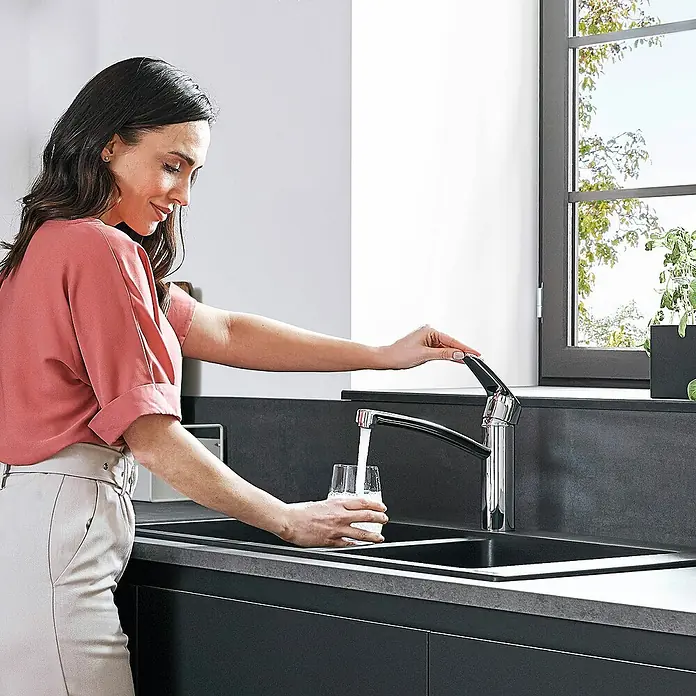 Hansgrohe Spültischarmatur Focus 160 - Chrom, Glänzend - Eine Frau befüllt ein Glas mit Wasser aus einem modernen Chrom-Wasserhahn in einer stilvollen, dunklen Küchenspüle.