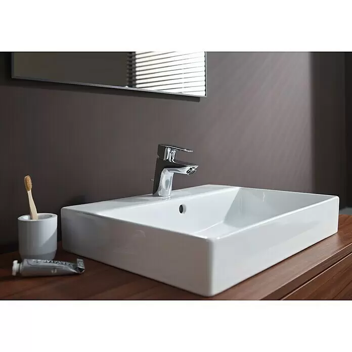 Hansgrohe MySport Waschtischarmatur M - Chrom, Glänzend - Rechteckiges weißes Keramikwaschbecken mit modernem Chromhahn auf einem Holzwaschtisch. Badezimmerutensilien und ein Spiegel mit Jalousien sind sichtbar.