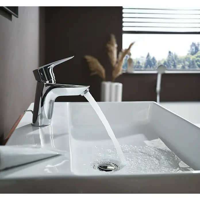 Hansgrohe MySport Waschtischarmatur M - Chrom, Glänzend - Nahaufnahme einer glänzenden Chromarmatur an einem weißen Waschbecken, aus der Wasser fließt. Im Hintergrund ein Fenster mit Blick in die Natur.