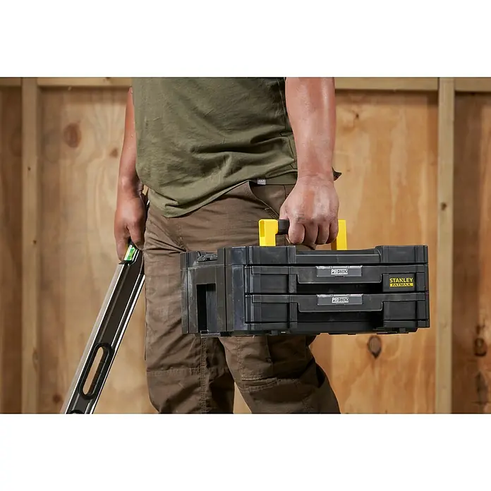 Stanley TSTAK Caja para herramientas con 2 cajones - 31,42 x 44 x 17,6 cm - Un artesano lleva una caja de herramientas negra Stanley Fatmax Pro-Stack con asa amarilla.