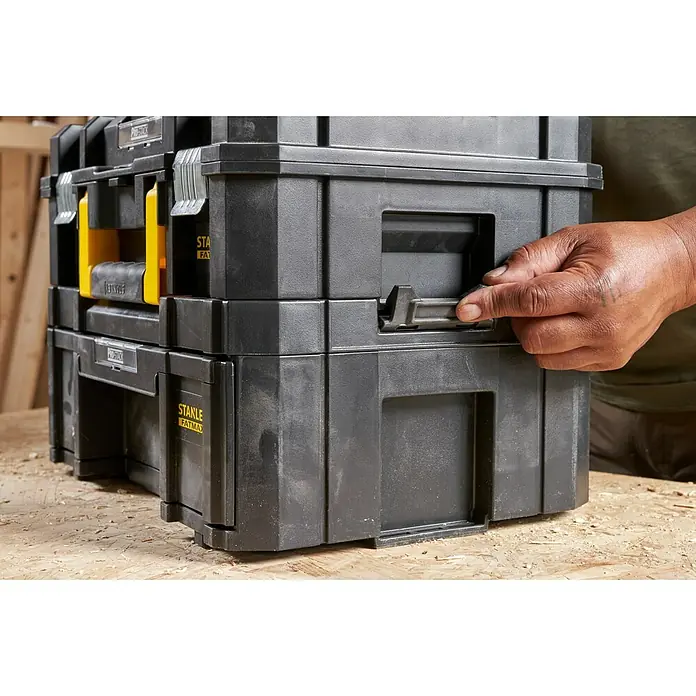 Stanley TSTAK Caja para herramientas Con cajones - 31,5 x 42,5 x 17,6 cm - Una pila de cajas de herramientas negras Stanley Fatmax con detalles en amarillo, con una mano accionando un mecanismo de bloqueo.