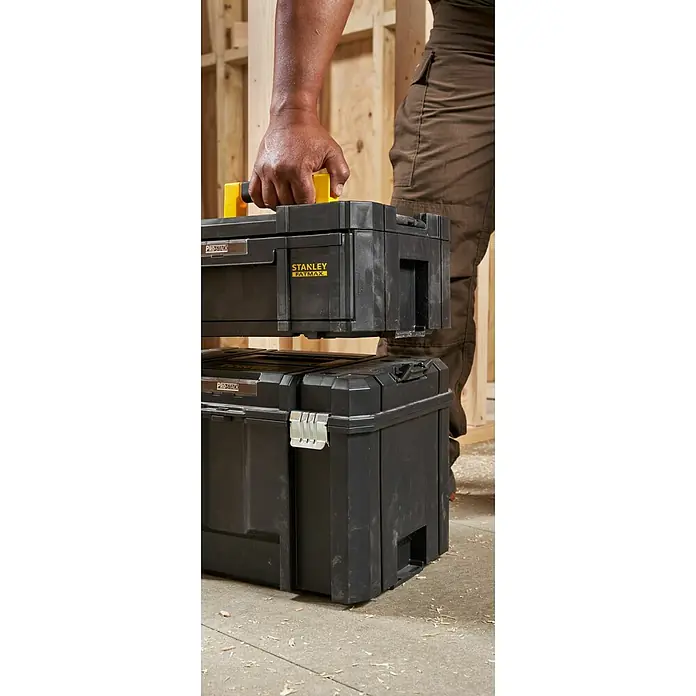Stanley TSTAK Caja para herramientas Con cajones - 31,5 x 42,5 x 17,6 cm - Una persona con ropa de trabajo lleva dos cajas de herramientas negras Stanley Fatmax Pro-Stack apiladas una encima de la otra.
