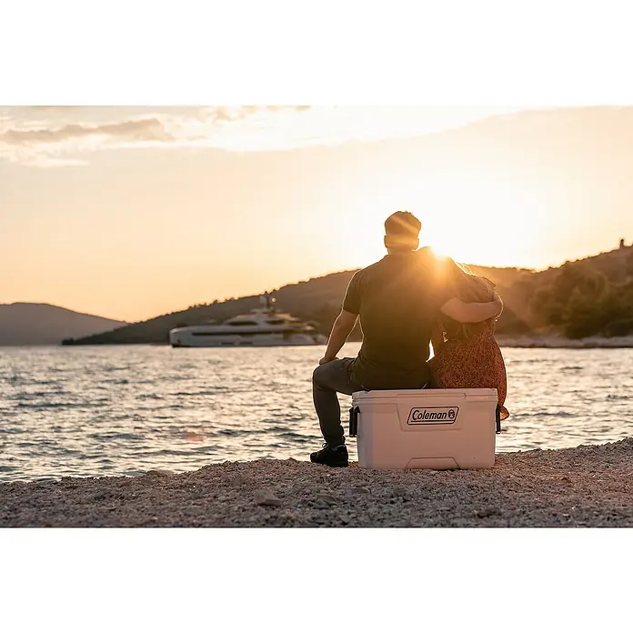Coleman Nevera de camping Xtreme Marine - L x An x Al: 39,5 x 79 x 43,5 cm, Blanco, 66 l - Una pareja sentada en una nevera Coleman en la playa, disfrutando de la puesta de sol con un yate al fondo.