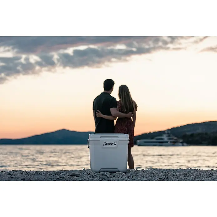 Coleman Nevera de camping Xtreme Marine - L x An x Al: 32 x 42 x 45 cm, Blanco, 26 l - Una nevera blanca de Coleman en la playa, mientras una pareja disfruta de la puesta de sol. Ideal para un picnic junto al mar.