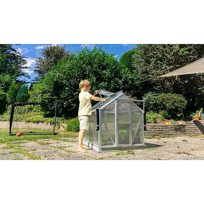 Vitavia Gewächshaus Merlin - Außenmaß inkl. Dachüberstand (B x T): 87,2 x 111,6 cm, Material Seitenwand: Kunststoff, 4 mm, Aluminium - Ein Junge öffnet das Dachfenster eines Gewächshauses im Garten, um die Pflanzen zu belüften. Das Gewächshaus ist aus Aluminium und durchsichtigem Kunststoff.