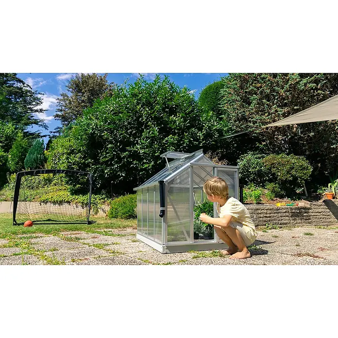 Vitavia Gewächshaus Merlin - Außenmaß inkl. Dachüberstand (B x T): 87,2 x 111,6 cm, Material Seitenwand: Kunststoff, 4 mm, Aluminium - Kleines Gewächshaus im Garten mit einem Kind davor. Das Gewächshaus ist aus Aluminium und hat transparente Wände, ideal für kleine Gärten.
