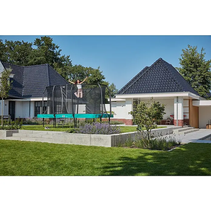 Salta Trampolin First ClassDurchmesser: 366 cm, Grün - Trampolin im Garten mit springendem Kind. Ein rechteckiges Salta Trampolin mit Sicherheitsnetz steht auf dem Rasen vor einem modernen Haus.