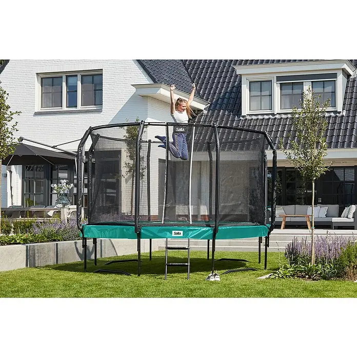 Salta Trampolin First ClassDurchmesser: 366 cm, Grün - Eine junge Frau springt auf einem Salta-Gartentrampolin mit Sicherheitsnetz vor einem Haus. Das Trampolin steht auf einem grünen Rasen.