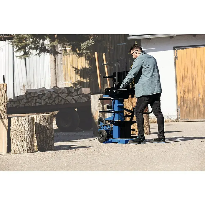 Scheppach Holzspalter Compact 15T - 15 t, Max. Spaltgutlänge: 107 cm - Ein Mann bedient einen blauen Holzspalter im Freien, um Holz zu verarbeiten. Holzscheite liegen im Hintergrund bereit.