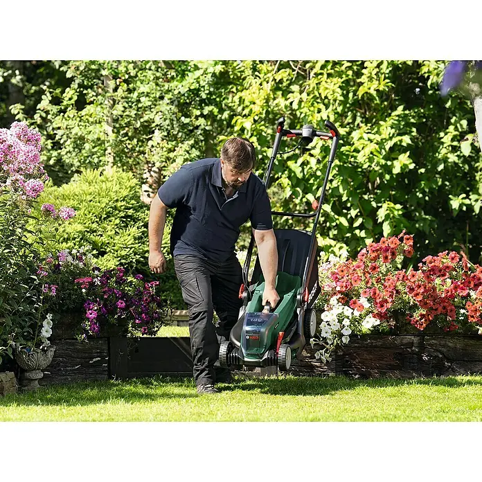 Bosch Power for All 18V Akku-Rasenmäher UniversalRotak 2x18V-37-550 - 18 V, Ohne Akku, Schnittbreite: 37 cm - Ein Mann schiebt einen grünen Bosch-Rasenmäher über einen Rasen mit Blumenbeeten. Der Rasenmäher ist das Hauptprodukt im Bild.