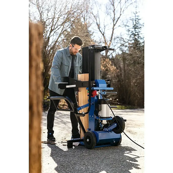 Scheppach Holzspalter Compact 15T - 15 t, Max. Spaltgutlänge: 107 cm - Ein Mann spaltet Holz mit einem blauen Holzspalter. Der Holzspalter ist mit Rädern ausgestattet und steht auf einer asphaltierten Fläche.