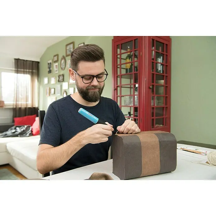 Bosch Akku-Heißklebestift Gluey - Farbe: Smoky Grey - Ein Mann arbeitet mit einem blauen Klebestift an einer braunen Box, im Hintergrund ein rotes Regal und helle Möbel.