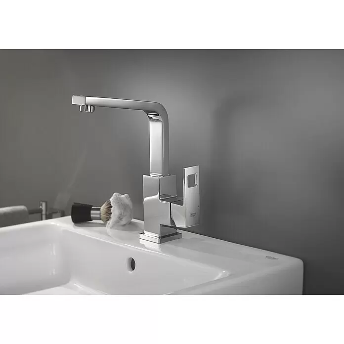 Grohe Eurocube Waschtischarmatur - Chrom, Glänzend, Schwenkbar - Polierter Chromarmatur im quadratischen Design auf einem weißen Waschbecken, mit einem Rasierpinsel daneben.