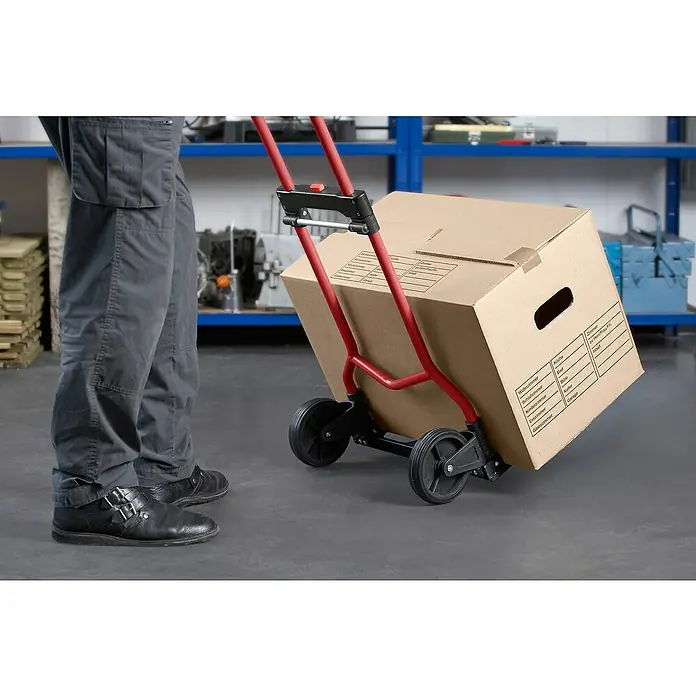 Carretilla de transporte plegable - Capacidad de carga: 60 kg, Aluminio - Un hombre transporta una caja de cartón con un salvaescaleras rojo. El salvaescaleras está equipado con ruedas negras y facilita el transporte.