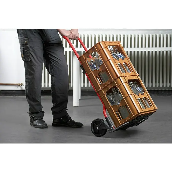 Carretilla de transporte plegable - Capacidad de carga: 60 kg, Aluminio - Una persona transporta dos cajas de botellas de bebidas en un carro rojo frente a un radiador.