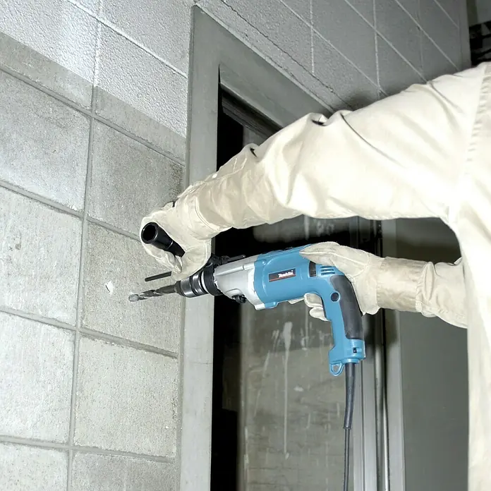 Makita Schlagbohrmaschine HP2071J - 1.100 W - Nahaufnahme einer Makita-Bohrmaschine, die in eine Betonwand bohrt, mit einer Person mit Handschuhen, die sie hält.