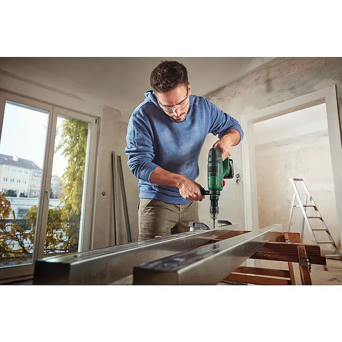 Bosch Professional AMPShare 18V Akku-Kombihammer UniversalHammer 18V - 18 V, 1 Akku, 4 Ah, 2 J - Ein Mann bohrt mit einer grünen Bohrmaschine ein Loch in ein Metallstück. Er trägt eine Schutzbrille.