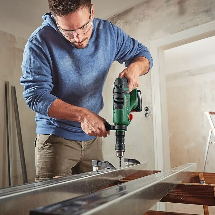 Bosch Power for All 18V Martillo combinado de batería UniversalHammer - 18 V, 1 batería, 4 Ah, 2 J - Un hombre perfora perfiles metálicos con un taladro percutor Bosch verde. Lleva gafas de seguridad y ropa de trabajo.