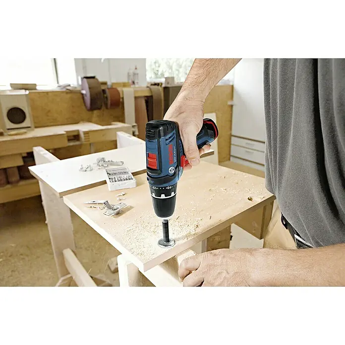 Bosch Professional Akku-Bohrschrauber GSR 12V-15 - 12 V, 2 Akkus, 2 Ah, 30 Nm, Ladegerät + Tasche - Akkuschrauber bei der Arbeit: Detailaufnahme von einem Bosch Akkubohrer beim Bohren in ein Holzbrett in einer Werkstatt.