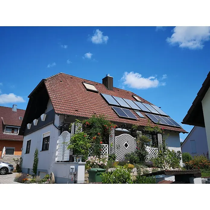 Sunset Dachhalterungs-Set - Passend für: Sunset Solaranlage Sunpay AS 300S - Ein Haus mit roten Dachziegeln und Solarmodulen auf dem Dach, umgeben von üppigem Grün und Blumen.