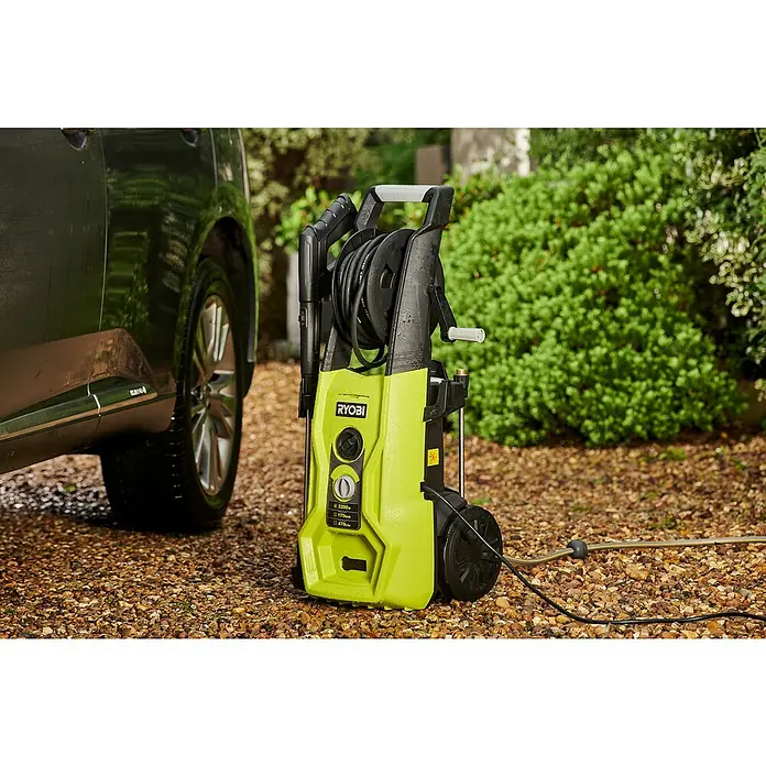Ryobi Hochdruckreiniger RY170PWA - 2.500 W - Druckreiniger der Marke Ryobi in Hellgrün und Schwarz neben einem dunklen Auto auf einem kiesbedeckten Untergrund. Das Gerät hat Räder und einen Griff.