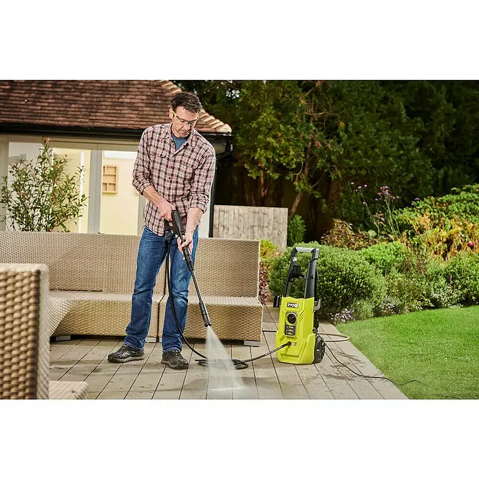 Ryobi Hochdruckreiniger RY130PWA - 1.600 W - Ein Mann benutzt einen Ryobi Hochdruckreiniger, um eine Holzterrasse zu reinigen. Der gelbe Reiniger steht rechts vom Mann auf der Terrasse.