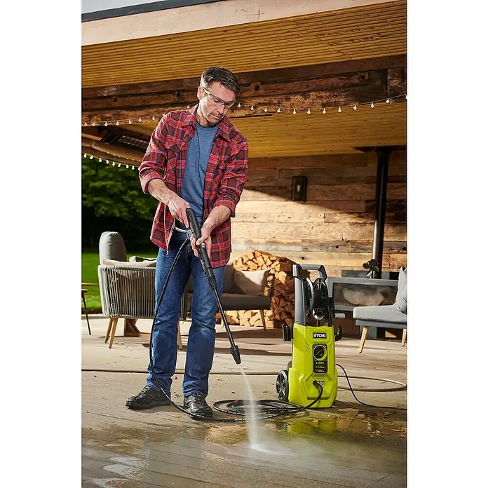 Ryobi Hochdruckreiniger RY140PWA - 1.800 W - Mann benutzt einen gelben RYOBI Hochdruckreiniger, um eine Terrasse zu reinigen, wobei er eine Schutzbrille und ein kariertes Hemd trägt.