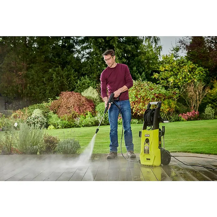Ryobi Hochdruckreiniger RY170PWA - 2.500 W - Mann reinigt eine Holzterrasse mit einem gelben Ryobi Hochdruckreiniger, Sicherheitsbrille tragend, umgeben von üppigem Grün.