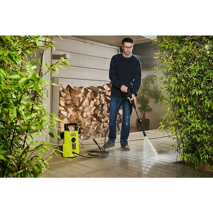 Ryobi Hochdruckreiniger RY110PWA - 1.400 W - Ein Mann benutzt einen gelben RYOBI-Hochdruckreiniger, um eine Holzterrasse zu reinigen. Das Gerät steht neben einem Holzstapel.