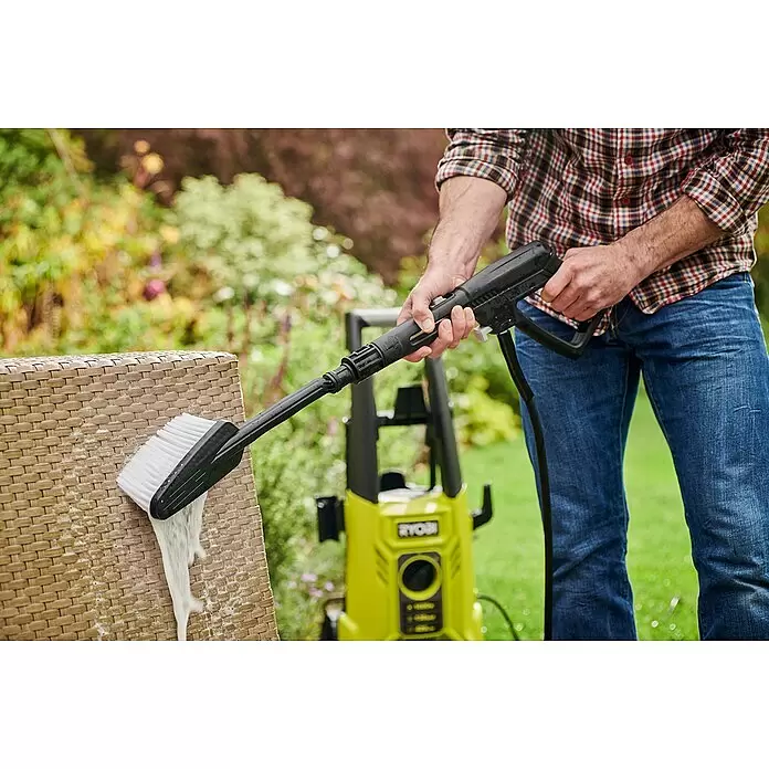 Ryobi Waschbürste RAC 774 - Passend für: Ryobi Hochdruckreiniger - Hände reinigen einen braunen Korbstuhl mit einer schwarzen Hochdruckreinigerbürste mit weißen Borsten, wobei Schaum entsteht. Ein grüner Hochdruckreiniger steht im Garten.