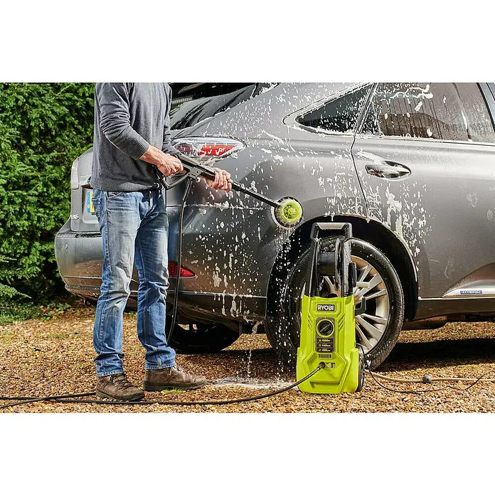 Ryobi Reinigungsbürste RAC 785 - Ausstattung: Borsten - Ein Mann wäscht ein graues Auto mit einem gelben Ryobi Hochdruckreiniger, der auf einem Kiesweg steht.