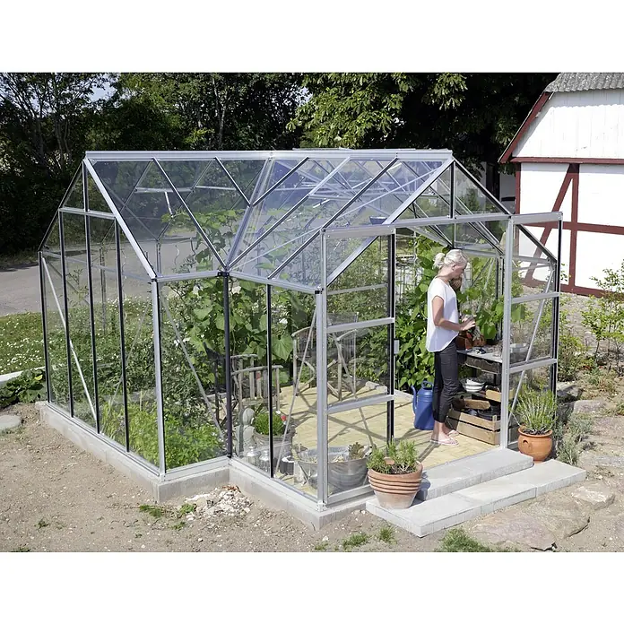Vitavia Gewächshaus Sirona - Außenmaß inkl. Dachüberstand (B x T): 383 x 383 cm, 3 mm, Aluminium - Gewächshaus mit einer Frau darin, die sich um die Pflanzen kümmert. Das Gewächshaus ist aus Glas und Metall gefertigt und beherbergt eine Vielzahl von Pflanzen.