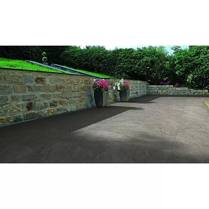 Terrassenfliese Manhattan 2.0 - 60 x 90 x 2 cm, Dunkelgrau, Matt - Außenbereich mit großen, dunkelgrau-braunen Steinoptikfliesen gepflastert, umgeben von Steinmauern und Grün.