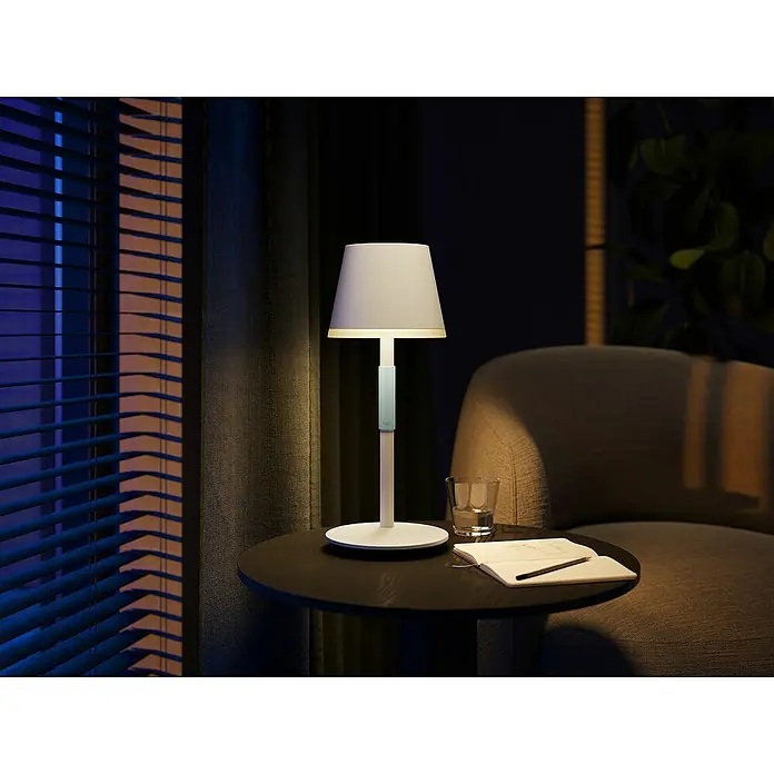 Philips Hue Akku-Tischleuchte Go Portable6,2 W, L x B x H: 21,9 x 20,9 x 38,8 cm, Weiß, Mehrfarbig - Weiße Tischlampe auf einem Tisch neben einem Sessel. Ein Glas Wasser und ein Notizbuch liegen ebenfalls auf dem Tisch.