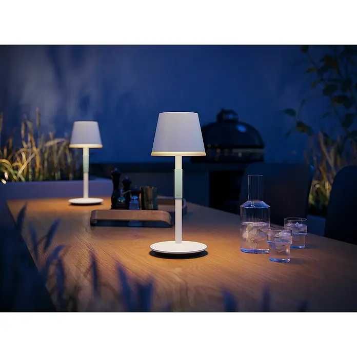 Philips Hue Akku-Tischleuchte Go Portable6,2 W, L x B x H: 21,9 x 20,9 x 38,8 cm, Weiß, Mehrfarbig - Ein stilvolles Ambiente mit zwei Tischleuchten, die warmes Licht auf einen Holztisch mit Getränken werfen.