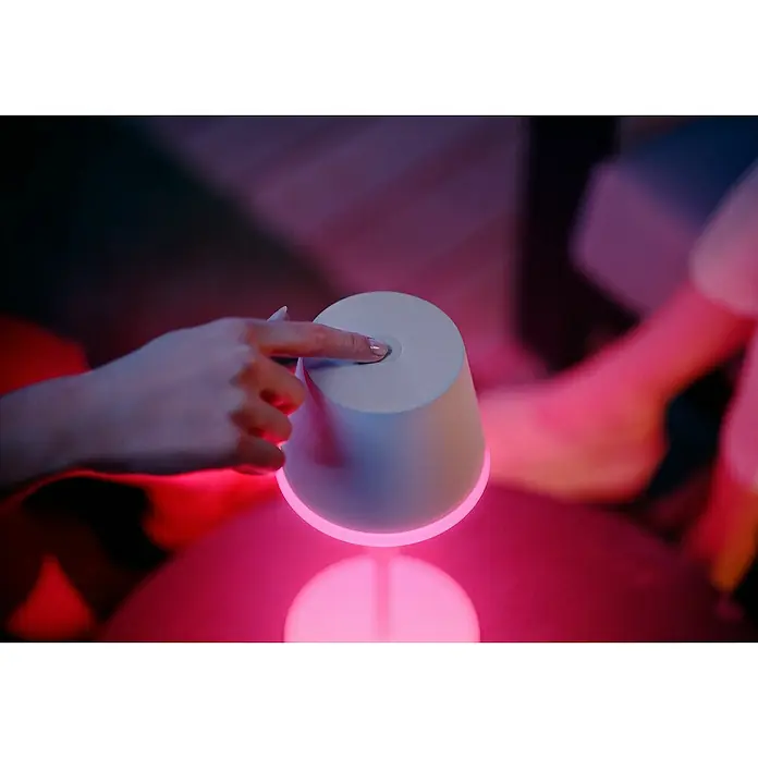 Philips Hue Akku-Tischleuchte Go Portable - 6,2 W, L x B x H: 21,9 x 20,9 x 38,8 cm, Mehrfarbig, Weiß - Nahaufnahme einer Hand, die eine moderne, farbwechselnde Tischleuchte mit sanftem, rosa Licht berührt.