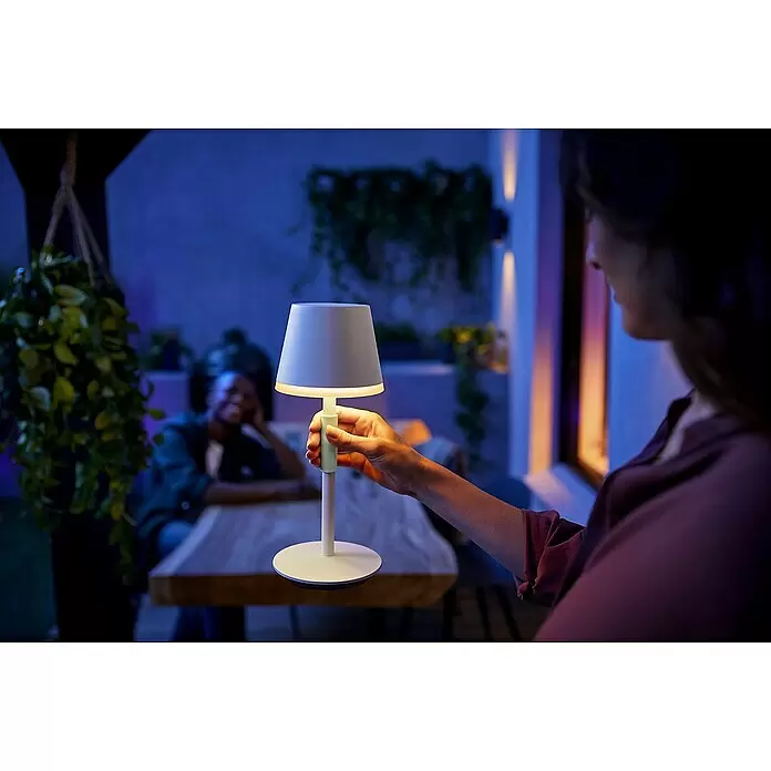 Philips Hue Akku-Tischleuchte Go Portable - 6,2 W, L x B x H: 21,9 x 20,9 x 38,8 cm, Mehrfarbig, Weiß - Eine Person hält eine weiße tragbare Tischlampe mit grünem Akzent, die in einer dunklen Umgebung warm leuchtet.