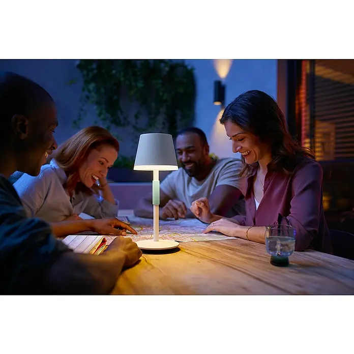 Philips Hue Akku-Tischleuchte Go Portable - 6,2 W, L x B x H: 21,9 x 20,9 x 38,8 cm, Mehrfarbig, Weiß - Stimmungsvolle Szene mit einer modernen Tischleuchte, die eine Gruppe von Menschen bei der Kartenplanung beleuchtet.