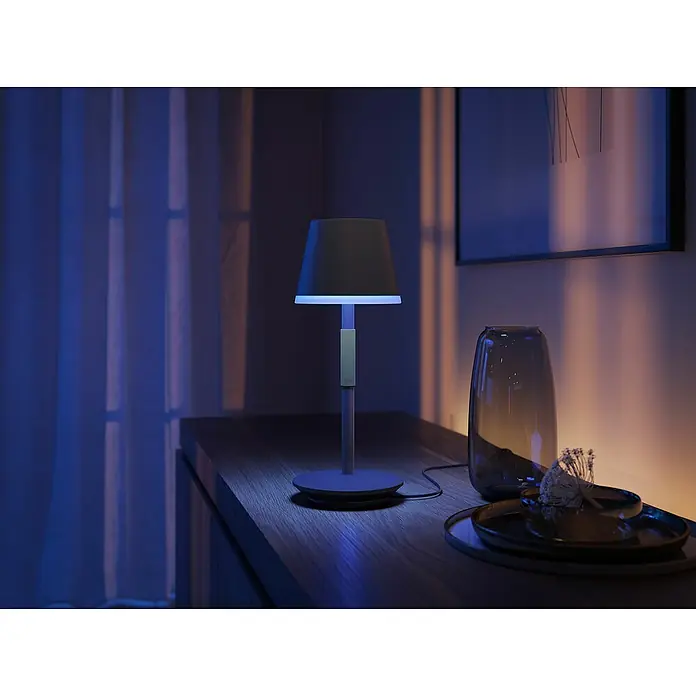 Philips Hue Akku-Tischleuchte Go Portable - 6,2 W, L x B x H: 21,9 x 20,9 x 38,8 cm, Mehrfarbig, Schwarz - Elegante Nachttischlampe mit blauem Licht auf einem Holztisch, neben einer Vase und Deko-Tellern, die eine warme Atmosphäre schafft.