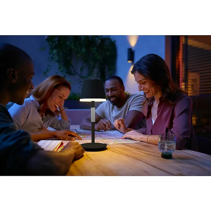 Philips Hue Akku-Tischleuchte Go Portable - 6,2 W, L x B x H: 21,9 x 20,9 x 38,8 cm, Mehrfarbig, Schwarz - Eine Tischlampe beleuchtet eine Gruppe von Freunden, die an einem Tisch sitzen und eine Karte betrachten.