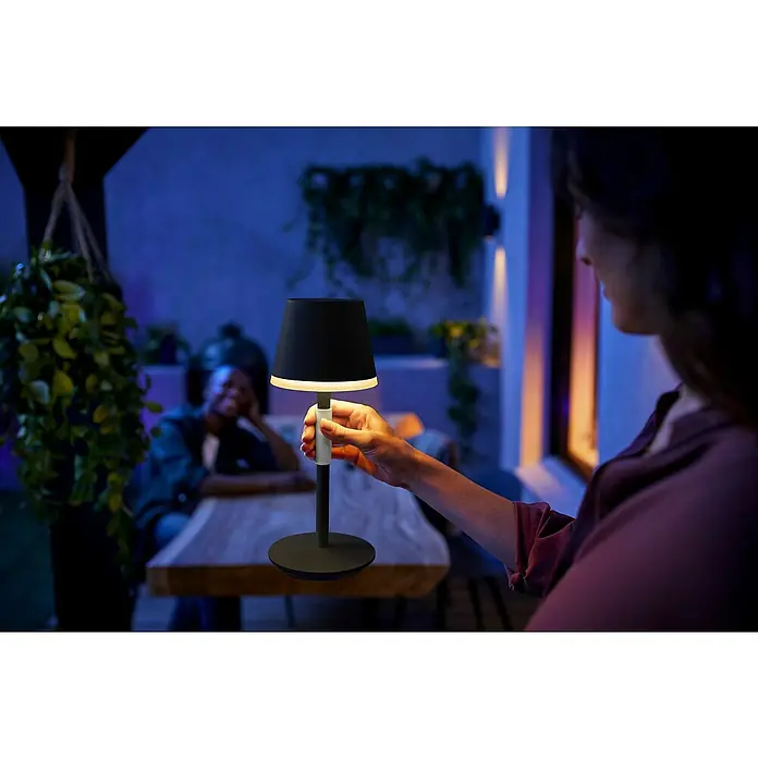 Philips Hue Akku-Tischleuchte Go Portable - 6,2 W, L x B x H: 21,9 x 20,9 x 38,8 cm, Mehrfarbig, Schwarz - Nahaufnahme einer kabellosen Philips Hue Tischleuchte mit schwarzem Lampenschirm, die von einer Person im Freien gehalten wird.