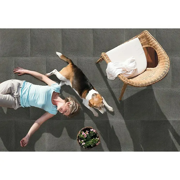 Terrassenfliese Manhattan 2.0 - 60 x 90 x 2 cm, Schwarz, Matt - Dunkelgraue Bodenfliesen auf einem sonnigen Hinterhof, wo eine Frau und ihr Hund sich entspannen. Der gewebte Stuhl und die Blumen vervollständigen die Szene.