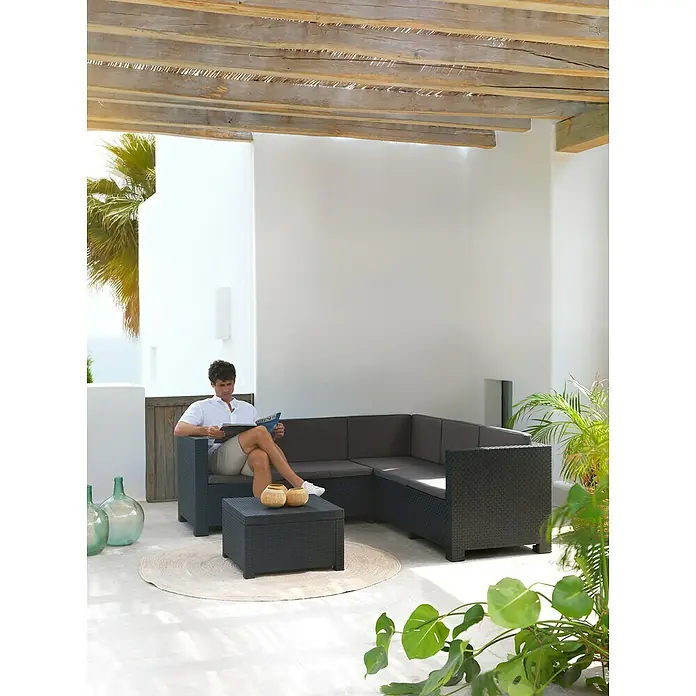 Shaf Conjunto de muebles Oasis - 3 pzs., Polipropileno, Grafito - Un hombre se relaja en un sofá seccional gris de exterior con cojines mientras lee una revista. El conjunto de asientos es de ratán.