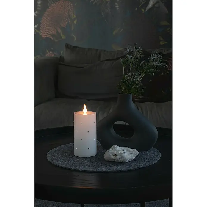 Konstsmide LED-KerzeWeiß, Höhe: 14,3 cm, 7,5 cm, Wachs - Stimmungsvolles Stillleben mit einer LED-Kerze mit Strasssteinen, einer modernen Vase mit Pflanzen und einem dekorativen Stein auf einem runden Tisch.