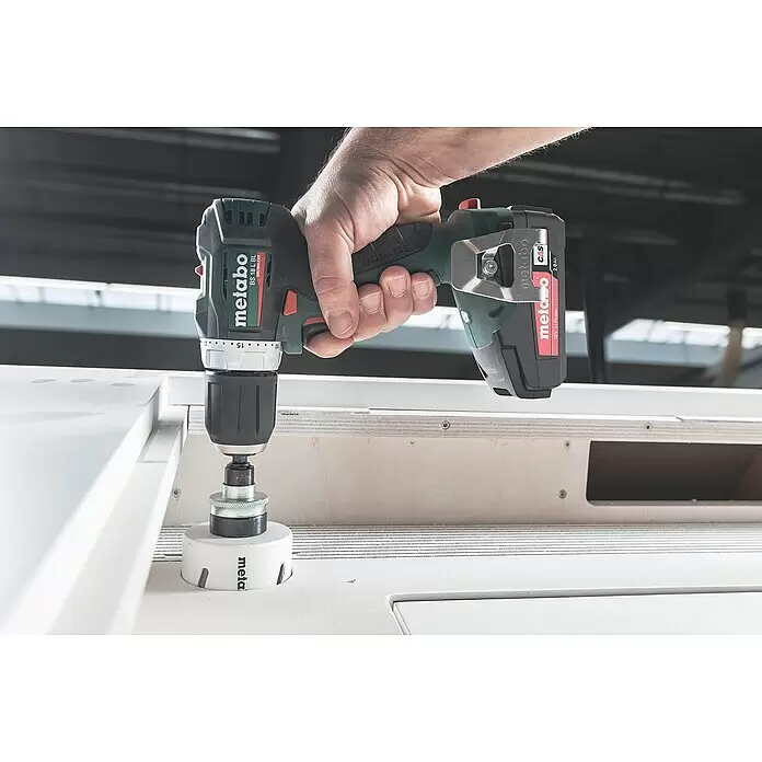 Metabo CAS 18V Akku-Bohrschrauber BS 18 L BL - 18 V, Ohne Akku, 60 Nm - Hand bohrt mit dunkelgrünem Metabo Akkuschrauber und weißer Lochsäge ein Loch in weißes Werkstück.