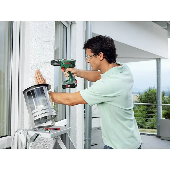 Bosch Taladro atornillador percutor de batería PSB 1800-Li - 18 V, 1 batería, 1,5 Ah, 38 Nm - Un hombre con gafas de seguridad utiliza un taladro inalámbrico Bosch para perforar una pared para instalar una lámpara.