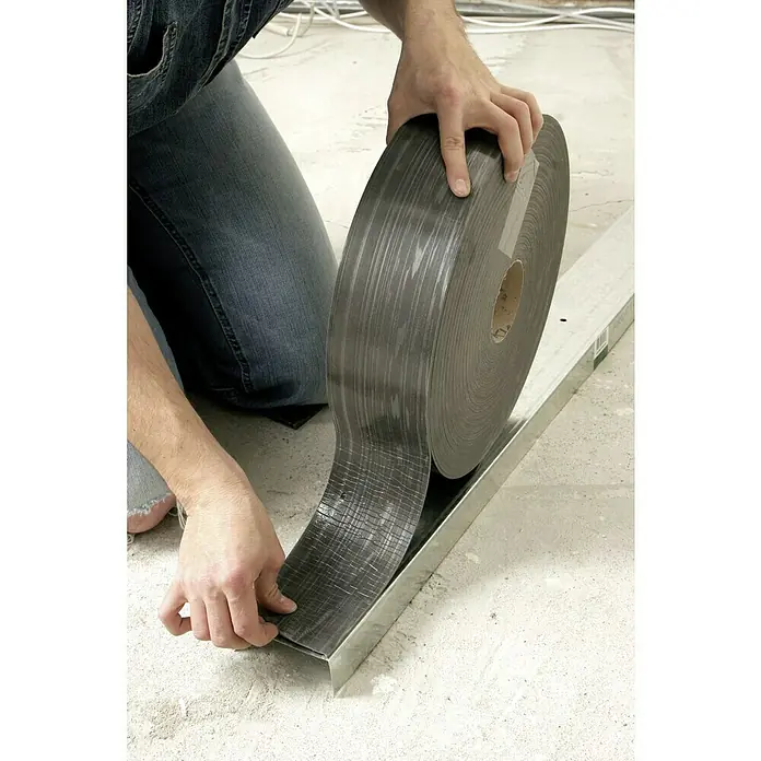 Knauf Dichtungsband - 10 m x 95 mm, Anthrazit - Eine Person verlegt eine Rolle graues Vinyl-Bodenbelag-Material in einen Metallrahmen, um einen sauberen Abschluss zu erzielen.