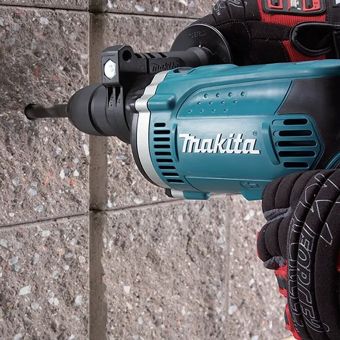 Makita Taladro percutor HP1631K - 710 W, Portabrocas de sujeción rápida - Primer plano de un taladro Makita perforando una pared de ladrillos, sujetado por una persona con guantes.