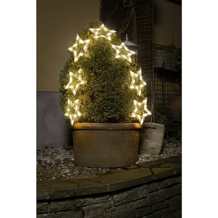 Konstsmide LED-Lichtervorhang Sternensilhouette - Außen, 10,72 m, Warmweiß, 70 Stk., Netzbetrieben - Dekorative Sternenlichterkette um einen grünen Busch in einem Terrakottatopf, die eine warme, festliche Atmosphäre erzeugt.