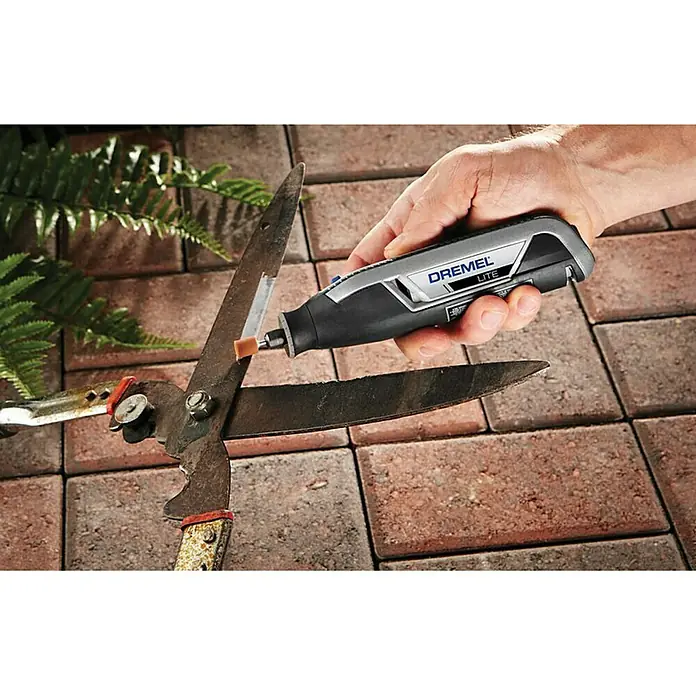 Dremel Akku-Multifunktionswerkzeug 7760-15 Lite - 3,6 V, 1 Akku, 2 Ah - Ein Dremel Lite wird verwendet, um eine große Gartenschere zu schärfen, die auf einem Pflastersteinweg mit etwas Grünzeug im Hintergrund liegt.